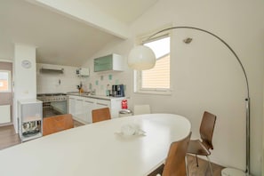 Et moderne køkken med hvide skabe, en komfur og et spiseområde med et hvidt bord og brune stole.