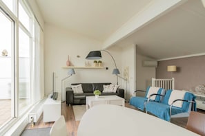 En moderne stue med en sofa, et sofabord og en blå lænestol. Der er et vindue til venstre, og en reol med pynteting over sofaen.