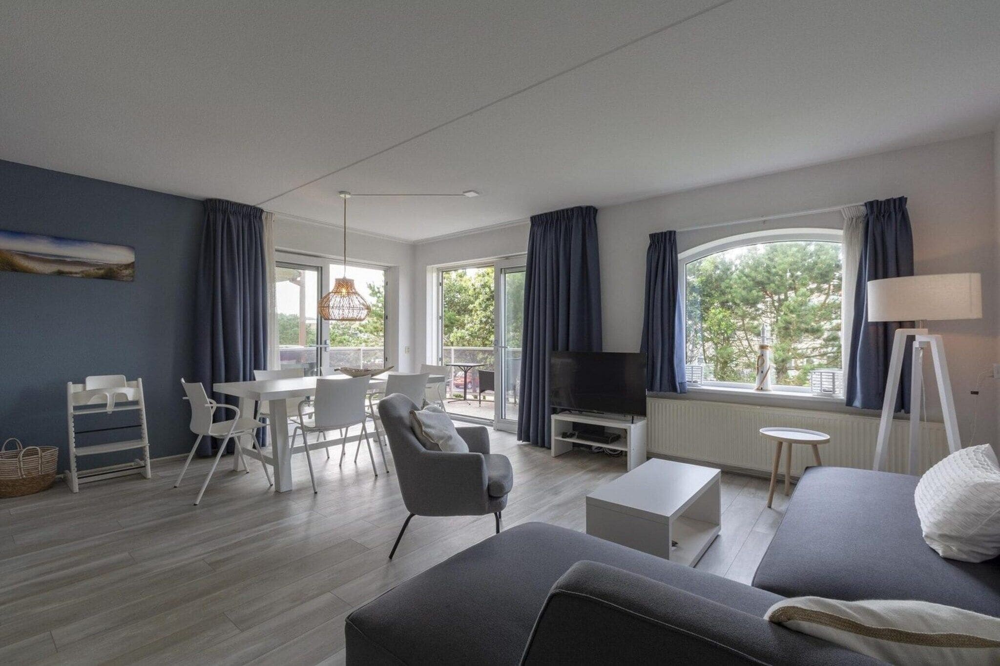 A modern living room with a sofa, a TV, a dining table with chairs, and a large window with curtains.