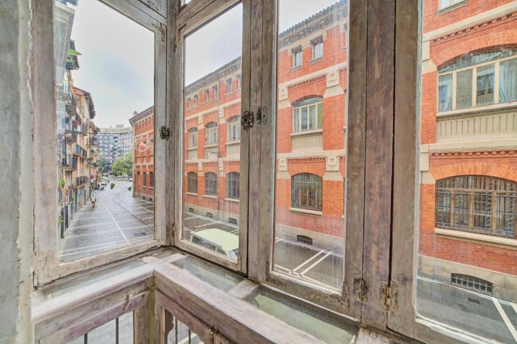 Vista a través de una ventana con marco de madera, que da a una calle con edificios de ladrillo rojo y cielo despejado.