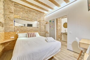 Un dormitorio con una cama grande, cabecera de madera, pared de piedra, techo de madera, una mesita de noche, un espejo y un baño visible a través de una puerta abierta.