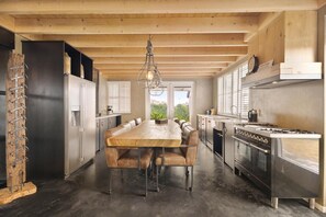 A modern kitchen with a large wooden table, stainless steel appliances, and a hanging light fixture. - Luxurious home behind the dunes of Julianadorp (Julianadorp)