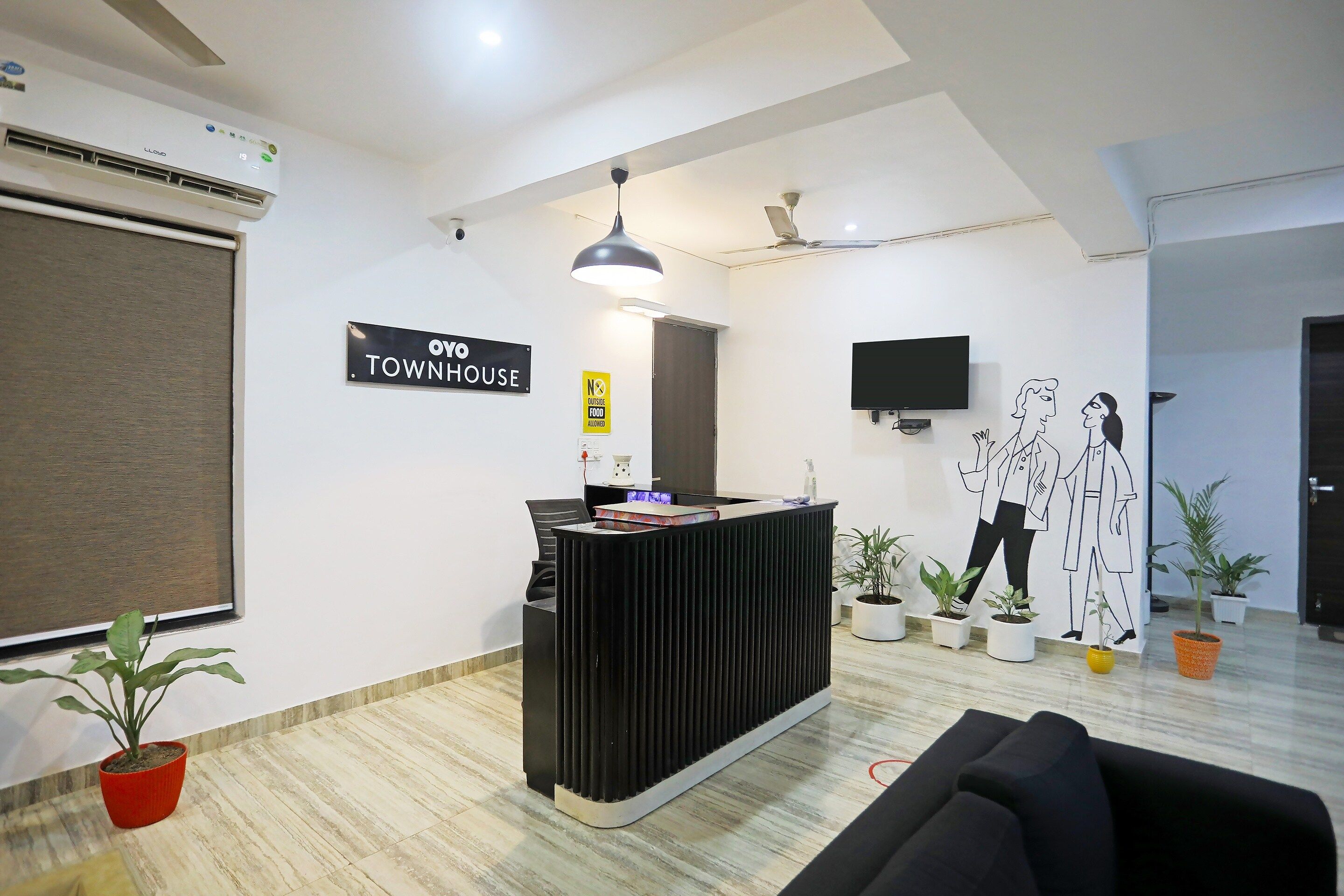 Una zona de recepció amb un taulell de recepció negre, un cartell que diu 'OYO TOWNHOUSE', un cartell amb la inscripció 'NO S'HI POTI PORTAR MENJAR DES DE FORA', un televisor de pantalla plana penjat a la paret i diverses plantes en testos.