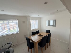 A dining area with a wooden table and black chairs, large windows with blinds, and a radiator on the wall. - House Sleeps 5 With Enclosed Garden (Corby)