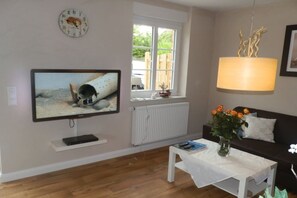 Ein Wohnzimmer mit Fernseher, Fenster, Heizkörper, weißem Tisch, einer Couch und einer Vase mit Blumen.