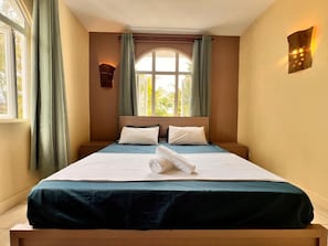 A hotel room with a bed, curtains, a window, and a wall-mounted light fixture. - Bazaar Square Lodges - Ylang-Ylang (Upper Floor) (Grand Baie)