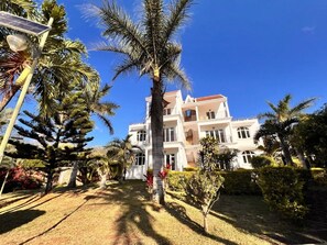 A multi-story building with a symmetrical facade, surrounded by palm trees and a well-maintained lawn. - Bazaar Square Lodges - Ylang-Ylang (Upper Floor) (Grand Baie)