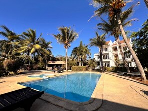 Ein Swimmingpool mit Liegestühlen, umgeben von Palmen und einem Gebäude mit weißer Fassade.