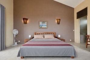 A bedroom with a bed, bedside lamps, a wall-mounted picture, and a floor fan. - Bazaar Square Lodges - Lotus (Panoramic Studio) (Grand Baie)