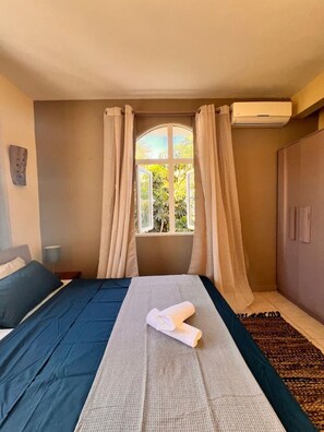 A bedroom with a bed, a window with curtains, a bedside lamp, and a wardrobe. - Bazaar Square Lodges - Hibiscus (Garden) (Grand Baie)