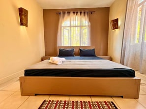 A bedroom with a large bed, a window with sheer curtains, and a patterned rug. - Bazaar Square Lodges - Frangipane (Garden) (Grand Baie)