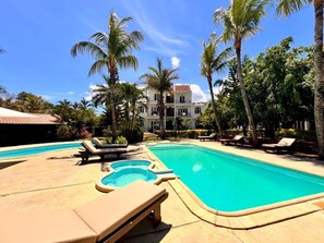 Zona de piscina con sillones de descanso, palmeras y un edificio al fondo.