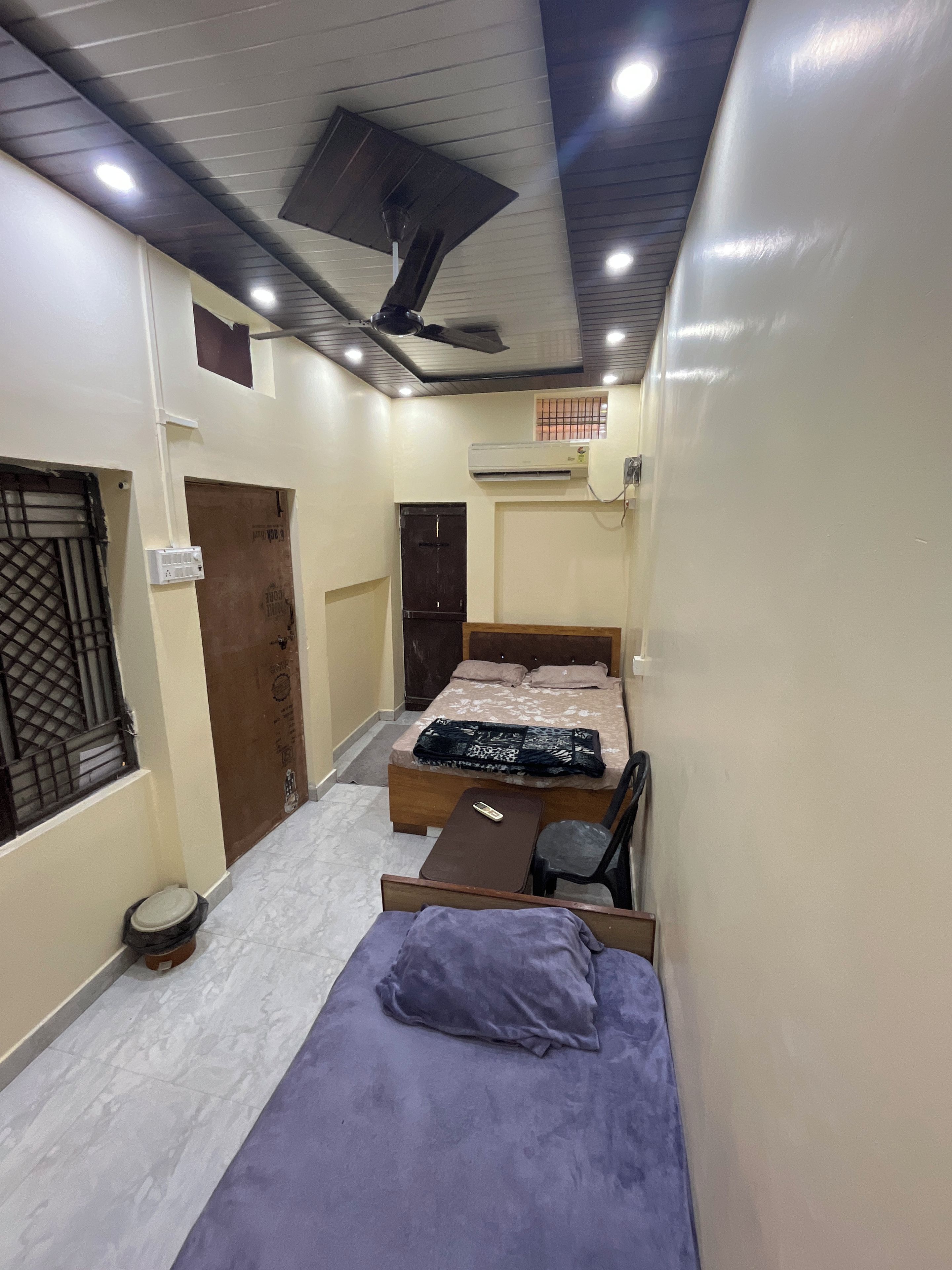 A compact room with a bed, a small table, a chair, an air conditioning unit, and a ceiling fan.