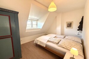 A bedroom with a sloped ceiling, a bed with patterned bedding, a bedside lamp, and a window. - "Reetstübchen" - House Shell (Nieblum)