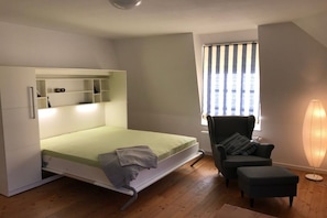 A compact room with a bed, a chair, a lamp, and a window with blinds. - "Villa Friedericia" - Apt. 04 (Wyk auf Föhr)