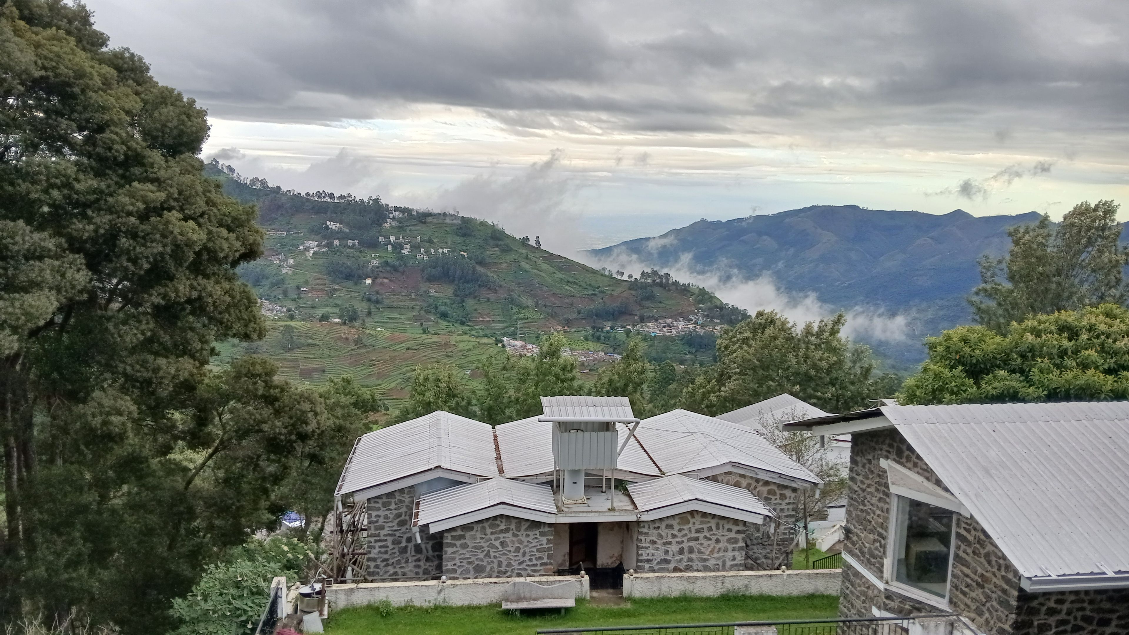 A stone building with a sloped roof, surrounded by lush greenery and a mountainous backdrop.