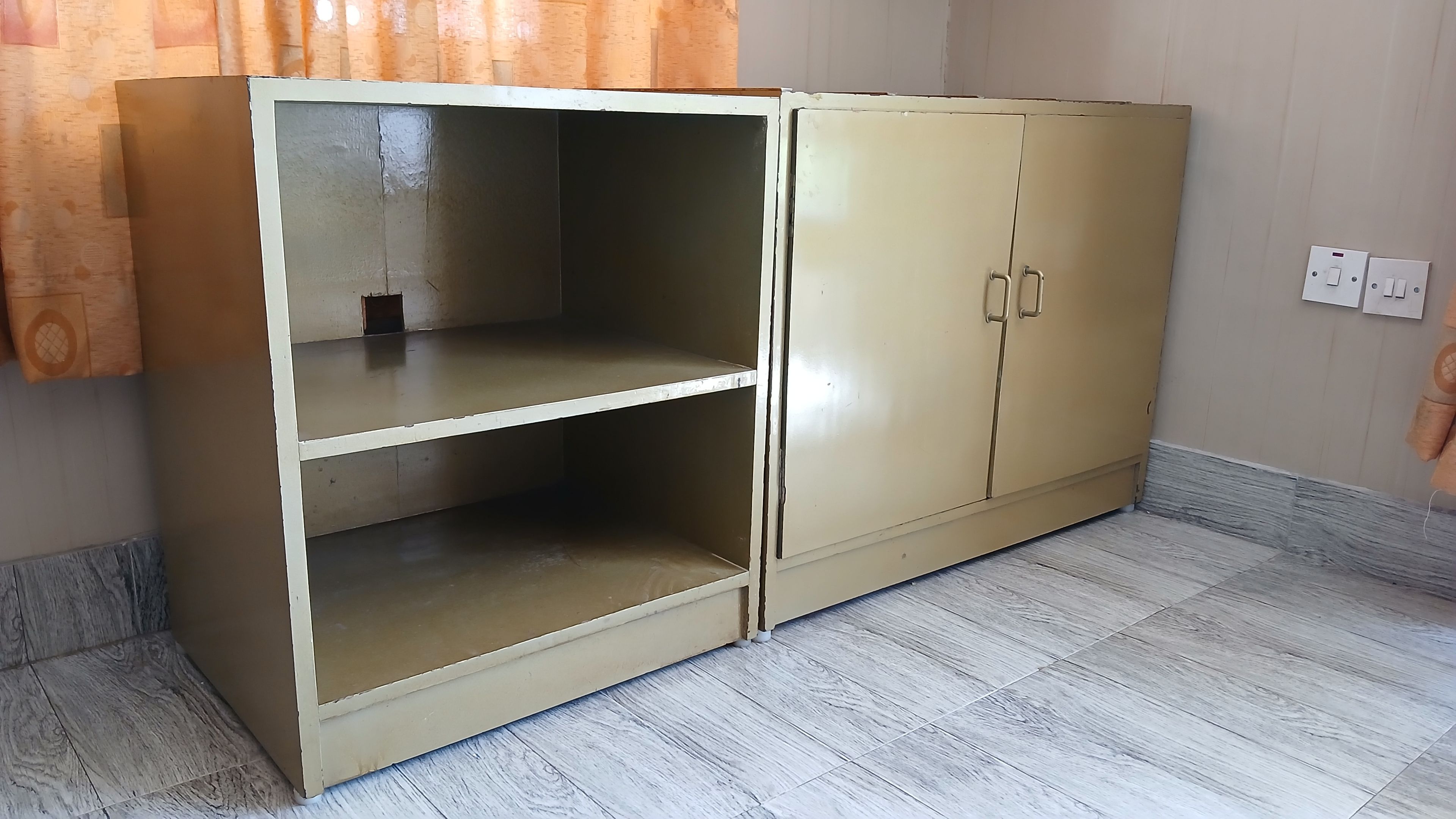 A beige wardrobe with two doors and two shelves, located in a room with light-colored wooden flooring and a curtain partially visible.