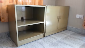 Un armario color beige con dos puertas y dos repisas, situado en una habitación con piso de madera claro y un cortin parcialmente visible.