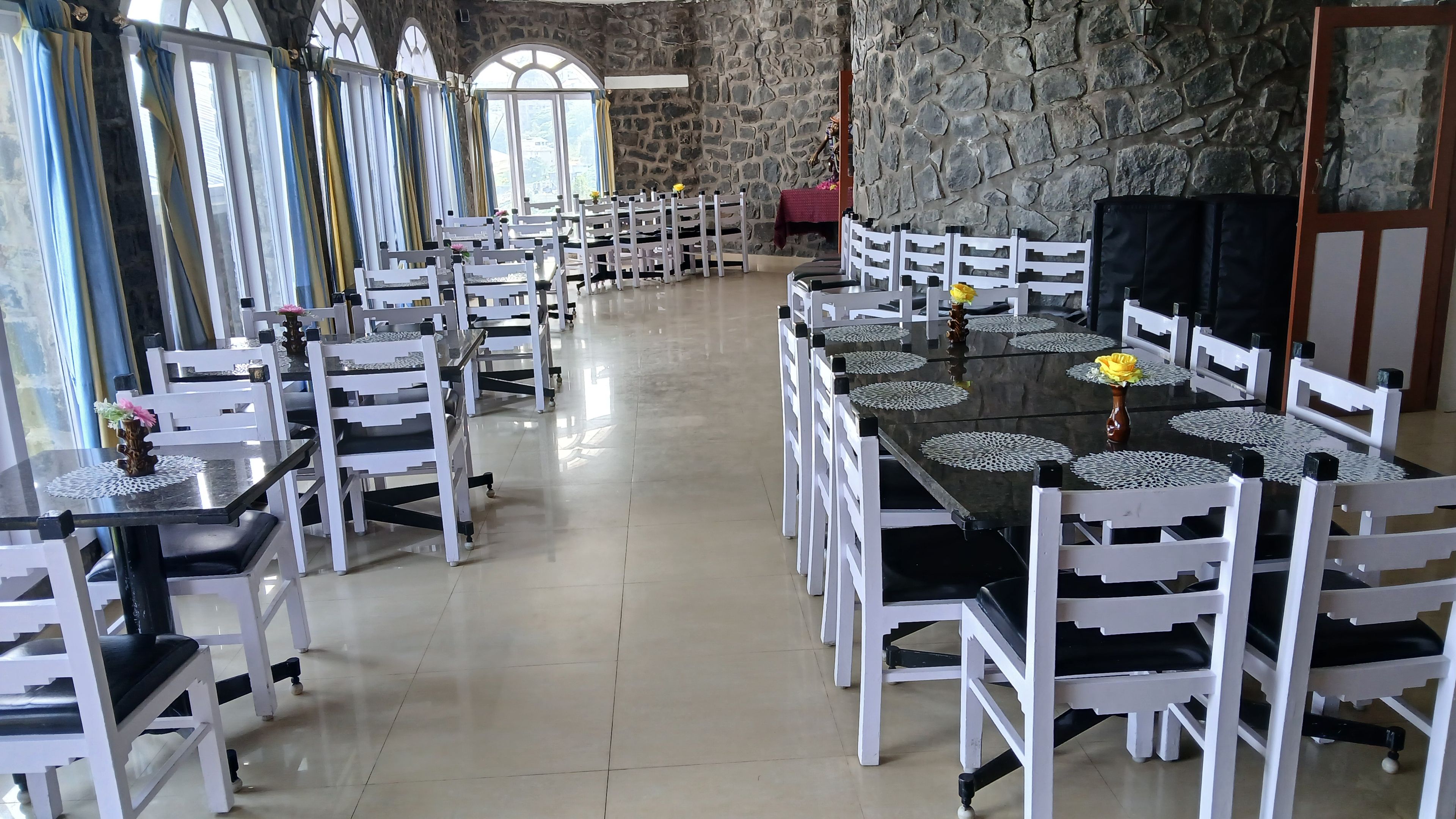 A spacious dining area with white chairs and black tables, stone walls, and large windows.
