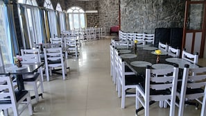 Un comedor amplio con sillas blancas y mesas negras, paredes de piedra y ventanas grandes.