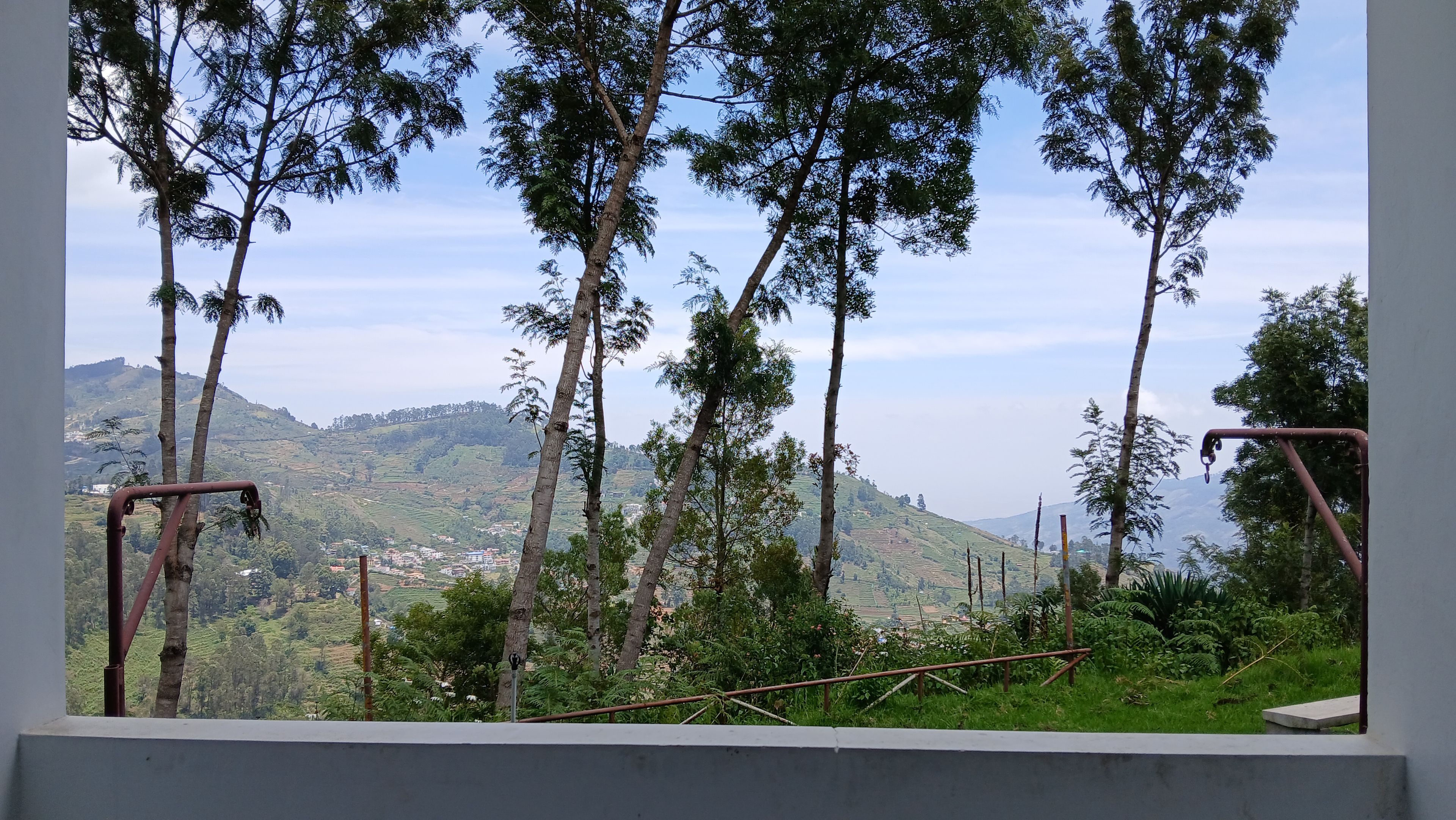 Vista a través de una ventana con baranda de metal, sobre un paisaje montañoso con árboles y un pueblo distante.