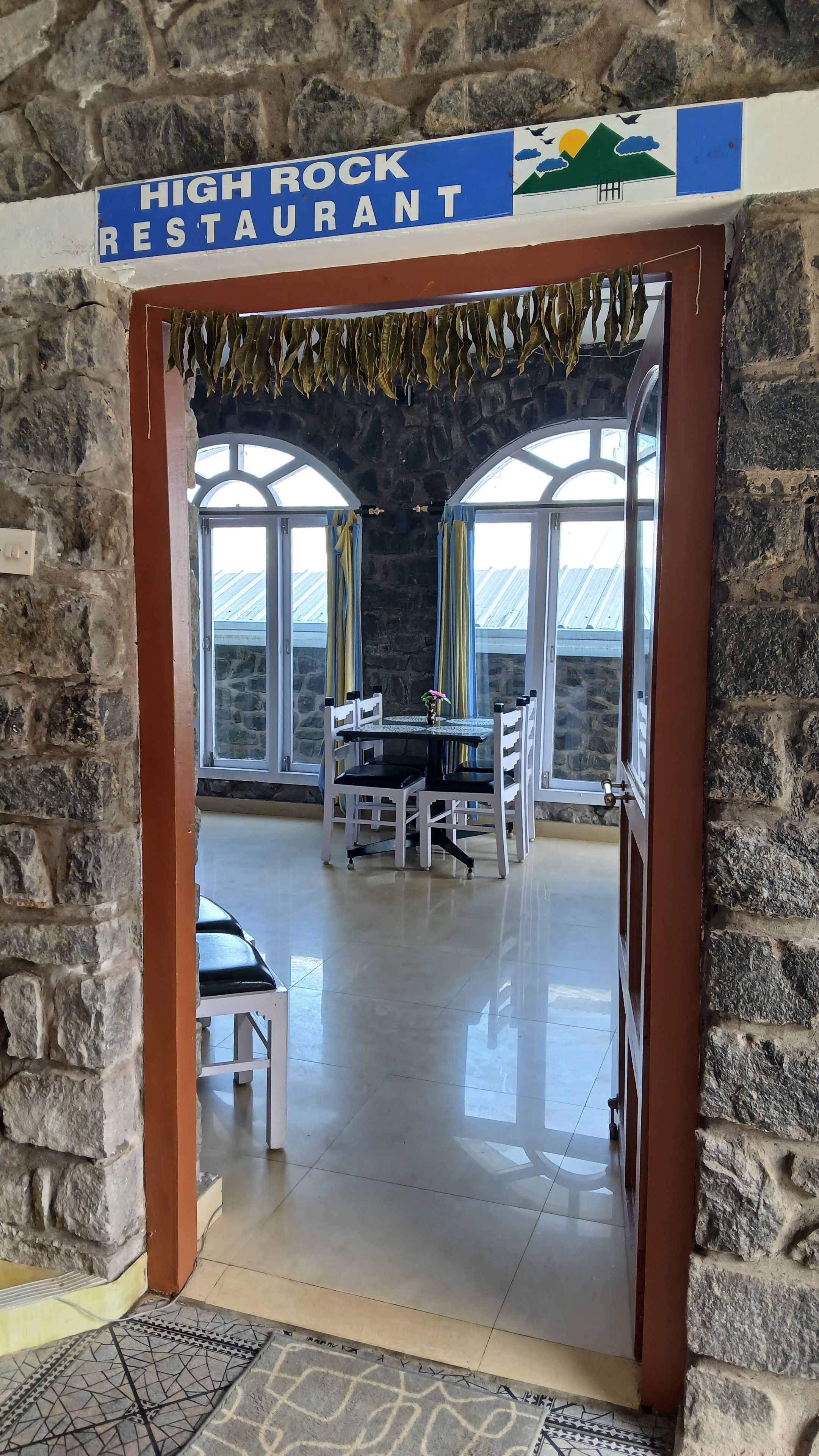 Entrada de um restaurante com parede de pedra, uma placa de identificação e área de jantar com mesas e cadeiras.