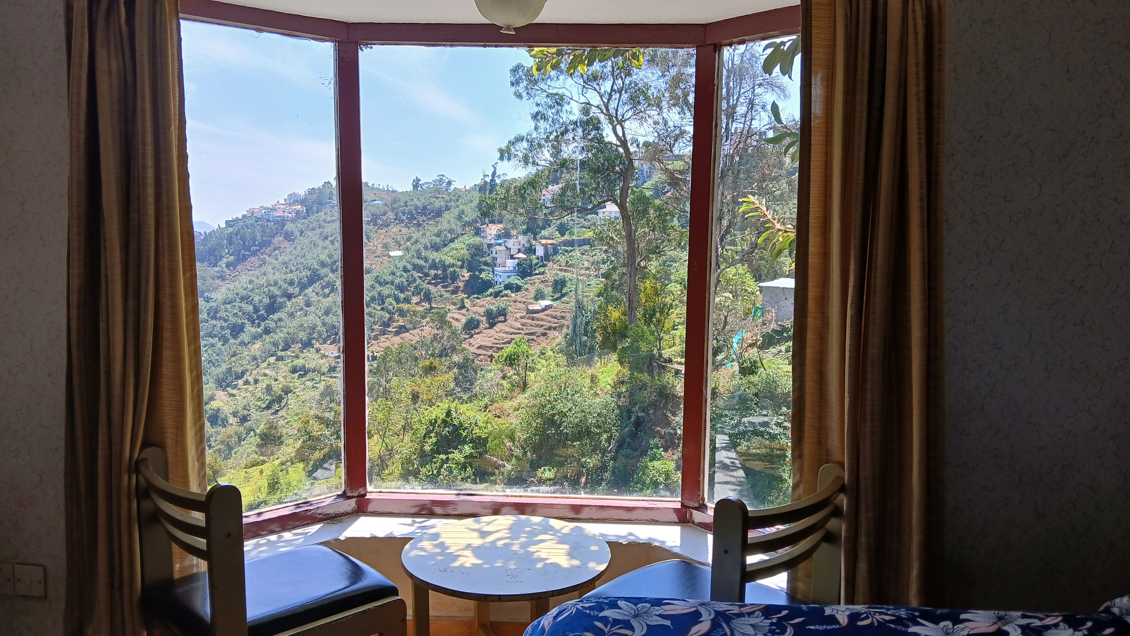 A room with a large window offering a view of a hilly landscape. There is a small table and two chairs in the room.