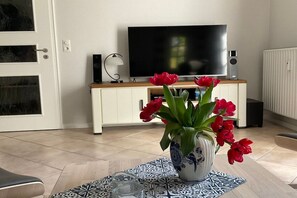 Un salon avec une télévision, des haut-parleurs, un vase rempli de fleurs rouges et une nappe à motifs.