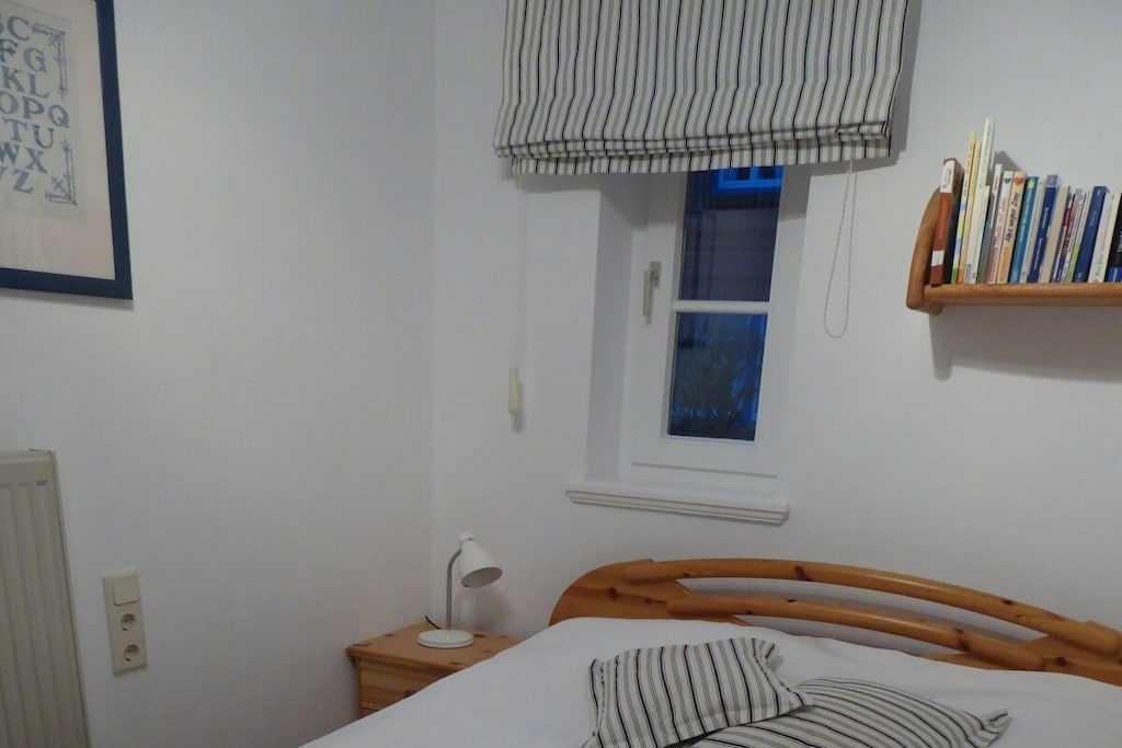 A bedroom with a wooden bed, a bedside table with a lamp, a window with a striped blind, and a bookshelf with books.