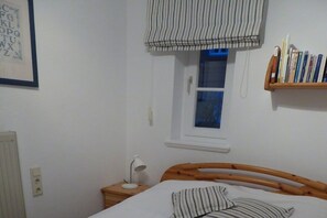 A bedroom with a wooden bed, a bedside table with a lamp, a window with a striped blind, and a bookshelf with books. - Holiday apartment with 1 bedroom (Wyk auf Föhr)