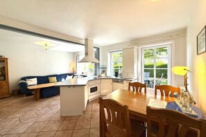 A kitchen with a central island, a dining table with chairs, a blue sofa, and a window with a view of greenery. - "Oldsum 2, House 94" (Oldsum)