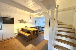 A kitchen with a wooden table and bench, a white staircase, and a living area with a sofa and a hanging light fixture. - Holiday apartment with 1 bedroom (Borgsum)