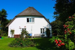 Une maison blanche avec un toit de chaume, entourée d’un jardin bien entretenu, agrémenté d’arbustes et d’un banc.