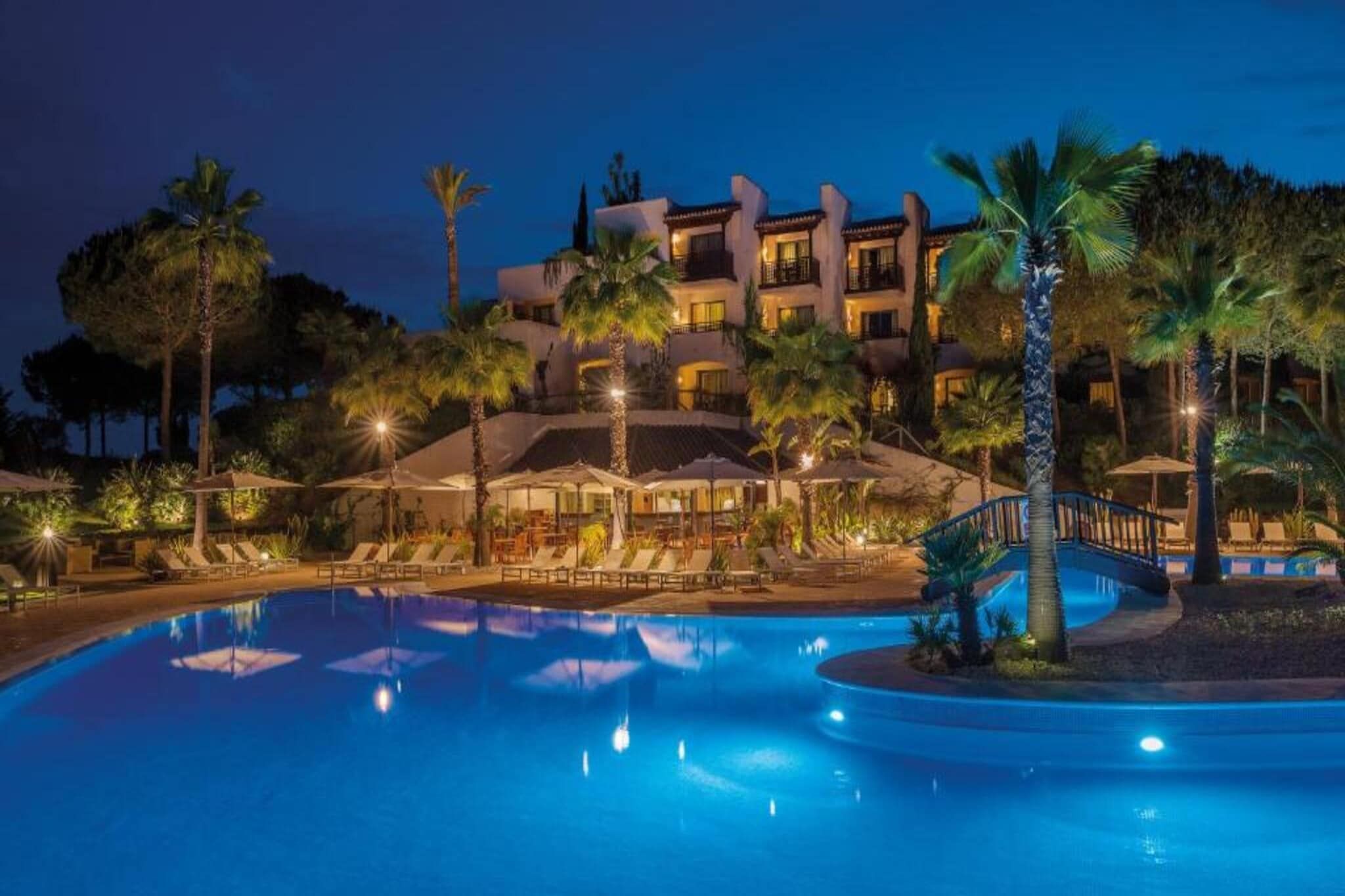 A nighttime view of a hotel with a swimming pool, palm trees, and outdoor seating areas.