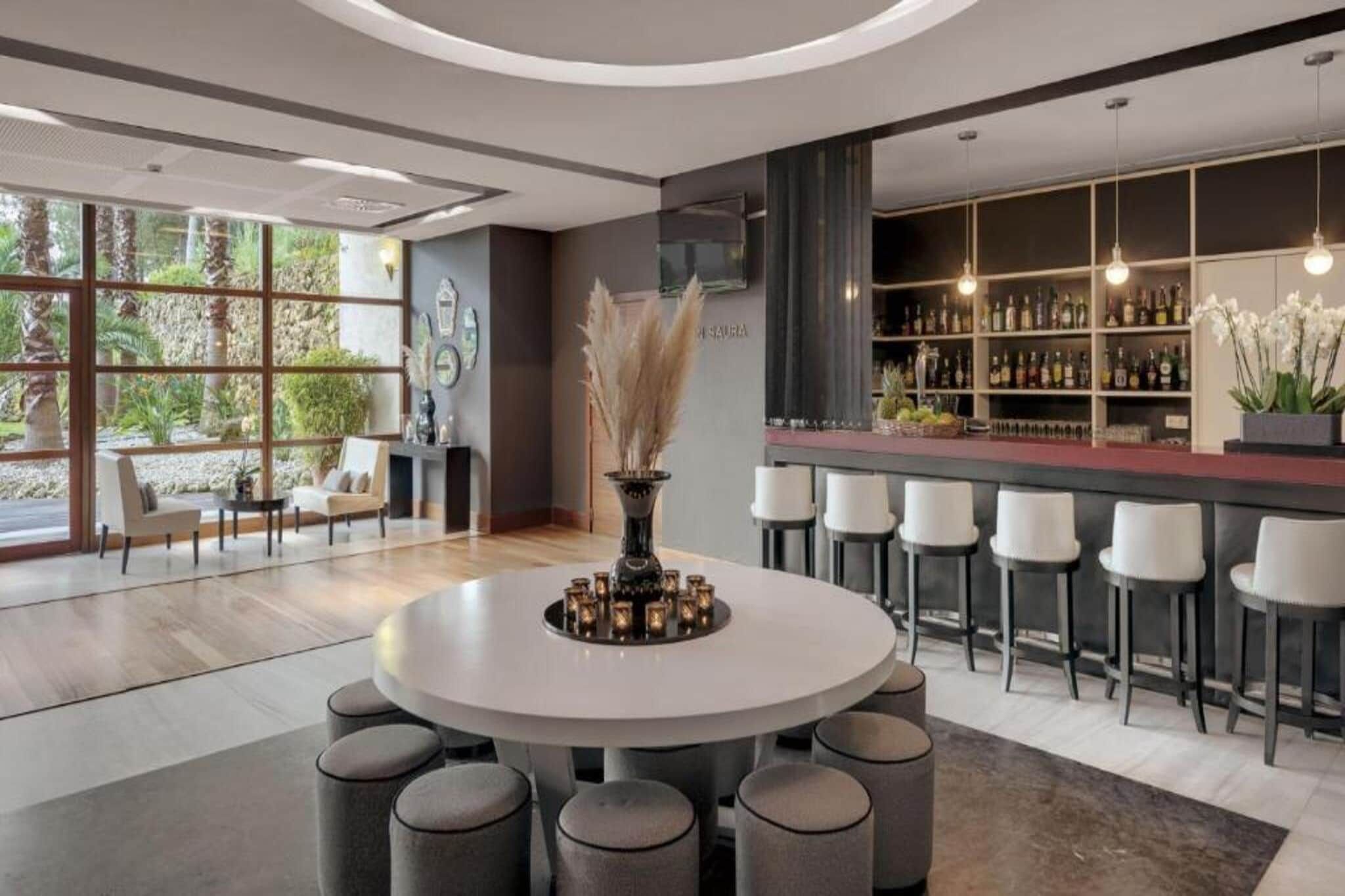 A modern interior with a round table surrounded by chairs, a bar area with shelves of bottles, and a view of greenery through large windows.