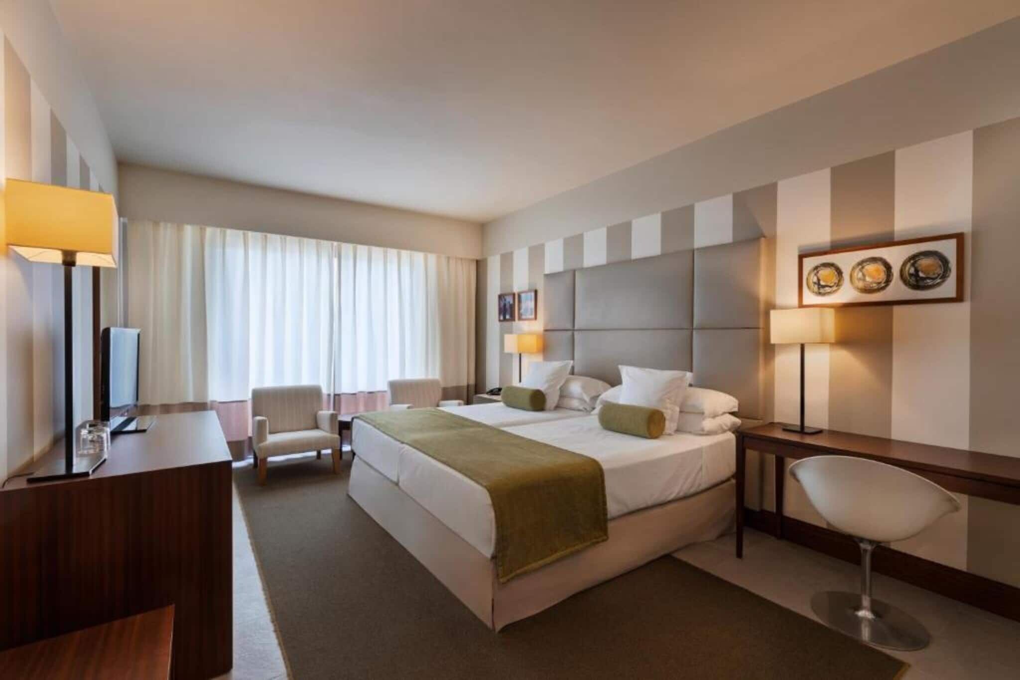 Habitación de hotel con cama, mesitas de noche, escritorio, silla, lámpara y ventana con cortinas.
