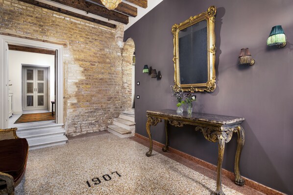 Une pièce avec un mur en briques, un grand miroir, une console ornée de vases et une porte donnant sur une autre pièce.
