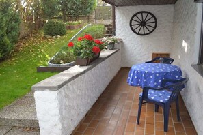Eine Terrasse mit Tisch und Stühlen, umgeben von einem Garten mit Kübelpflanzen und einer Wanddekoration in Form eines Rades.