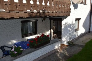 A white building with a wooden lattice balcony, a bench with flowers, and a door leading to an interior space. - Holiday apartment with 1 bedroom (Hopferau)