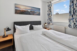 Ein Schlafzimmer mit einem Bett, einem Nachttisch, einer Lampe, einem Fenster mit Vorhängen und einem Bild an der Wand.