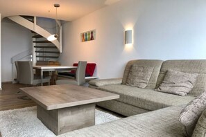 A modern living room with a sofa, a coffee table, a dining table with chairs, a staircase, and a wall-mounted light fixture. - Penthouse Swallow's Nest (Damp)