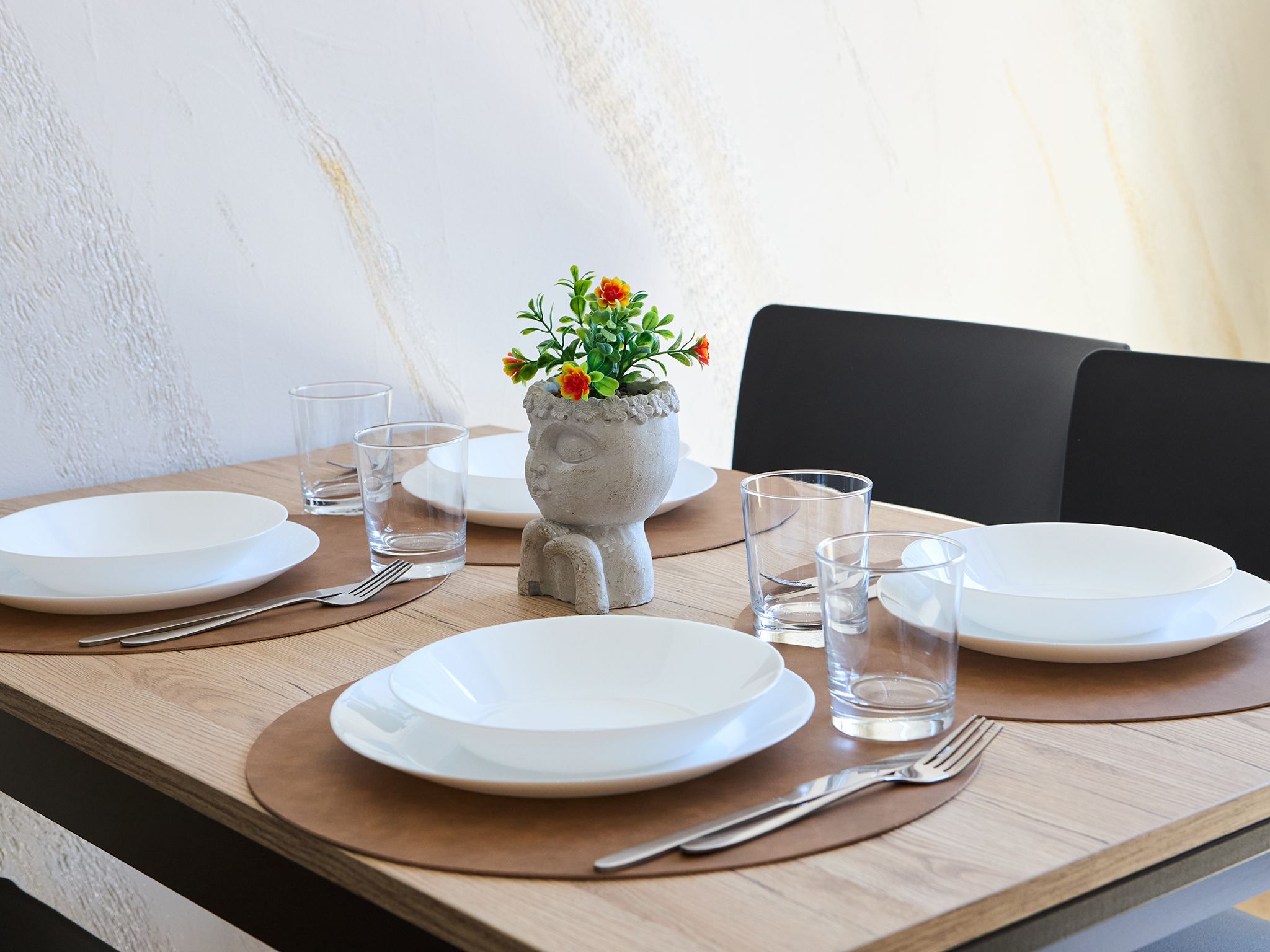 A dining table set with white plates, clear glasses, and cutlery, featuring a unique stone vase with flowers.