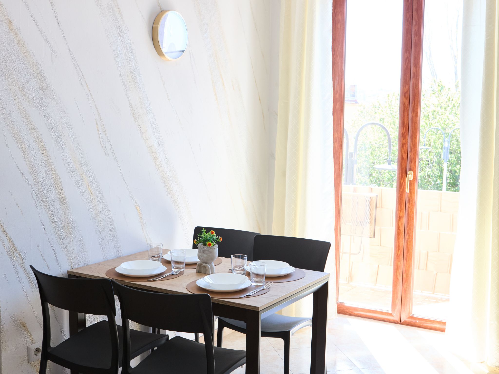 A dining area with a table set for four, a vase with flowers, and a view of greenery through a glass door.