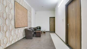 Un pasillo con baldosas de diseño, una zona de estar con sillones y pufs de cuero, una mesa con superficie de vidrio y una puerta de madera con un número.