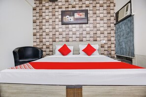 Habitación de hotel con pared de madera con diseño a cuadros, cama con ropa blanca y almohadas rojas, una butaca de cuero negro y un cuadro en la pared.