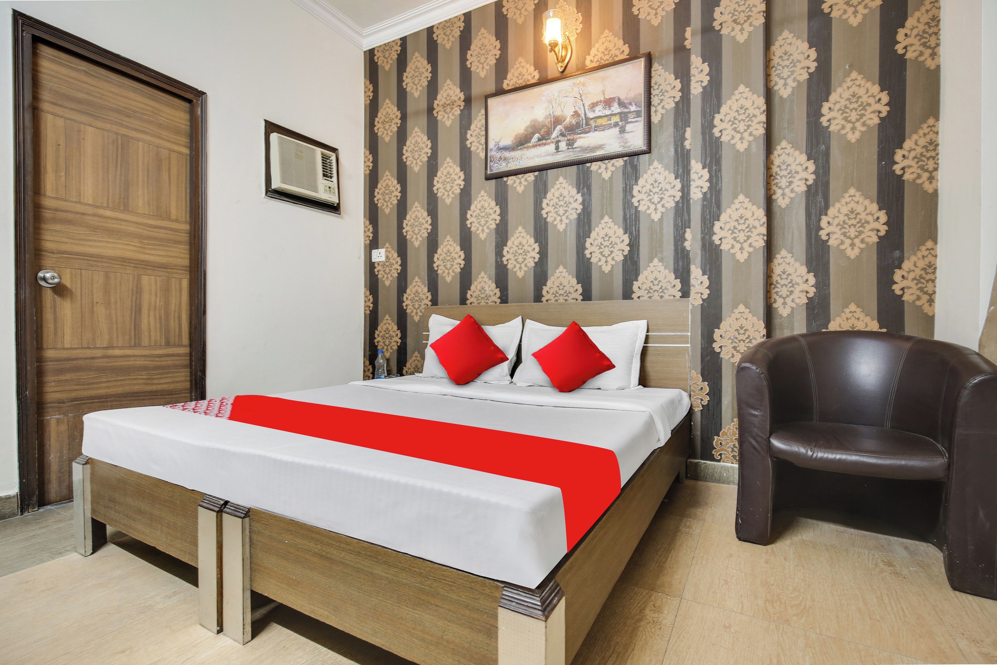 Een hotelkamer met een houten bed, een rood-wit beddekleed, een rood-wit kussen, een bruine leren fauteuil en behang met een patroon.