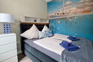A bedroom with a bed, pillows, a nightstand, a lamp, and a mural of a ship on the wall. - 2 - House Windröschen (Borkum)
