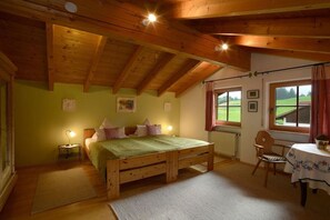 A room with a wooden bed, a table, and a chair. There are windows with curtains, a radiator, and a picture on the wall. - Ellen Raith - Apartment 2 (Füssen)