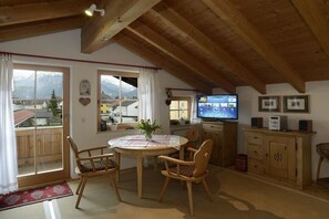 A cozy room with a wooden ceiling, a dining table with chairs, a television, and a view of mountains through large windows. - Ellen Raith - Apartment 2 (Füssen)