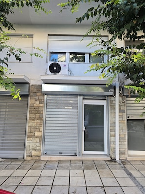 A building with a closed metal shutter door, a stone wall, and an air conditioning unit on the roof. - Modern apartment in Thessaloniki (Θεσσαλονίκη)